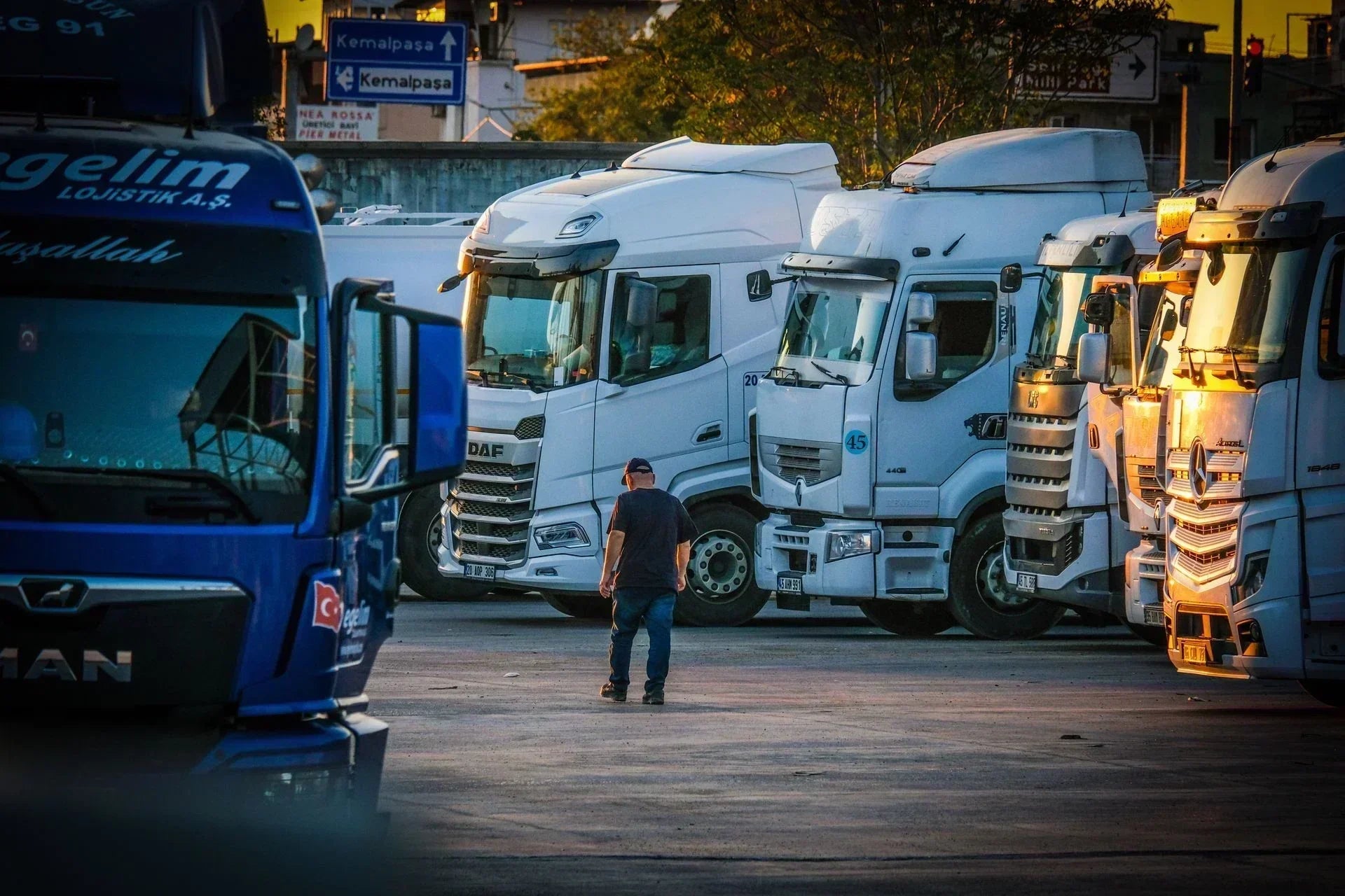 A professional trucker walking through a busy truck stop parking lot surrounded by heavy-duty commercial rigs and transport trailers.